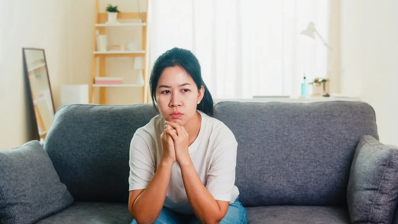 An Asian woman, depressed and crying, stressed with a headache, sits on a sofa in the living room of a house.